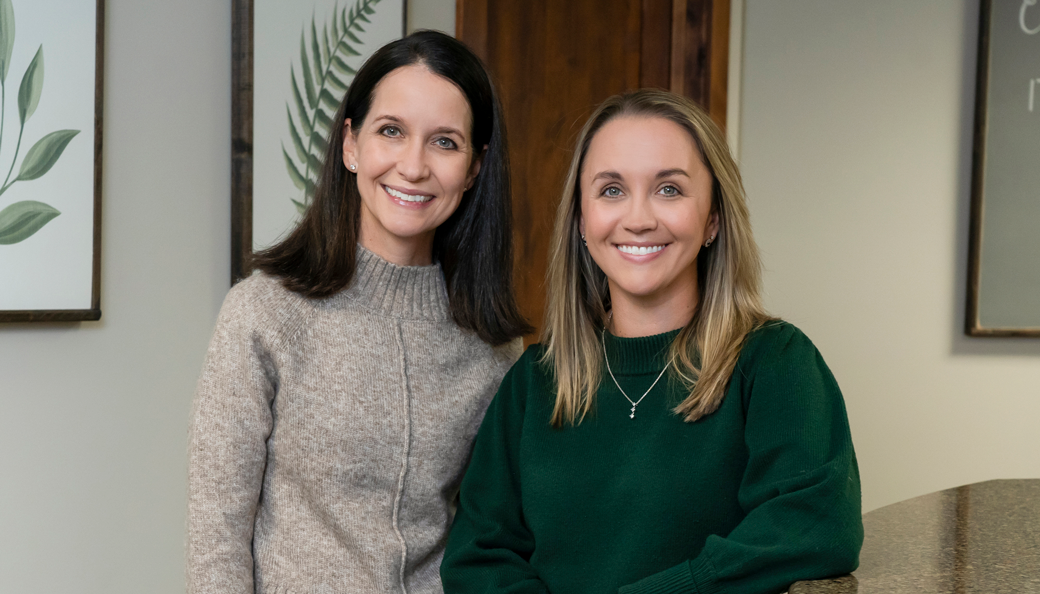 Platt Dentistry | Two women smiling in a warmly lit office setting, one wearing a beige knit sweater and the other in a dark green sweater, standing in front of framed botanical prints.